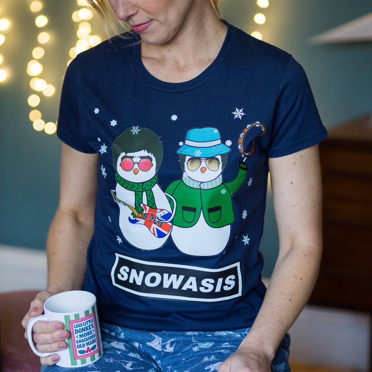 Person wearing a navy blue Christmas t-shirt with cartoon snowmen and 'SNOWASIS' text, holding a mug.