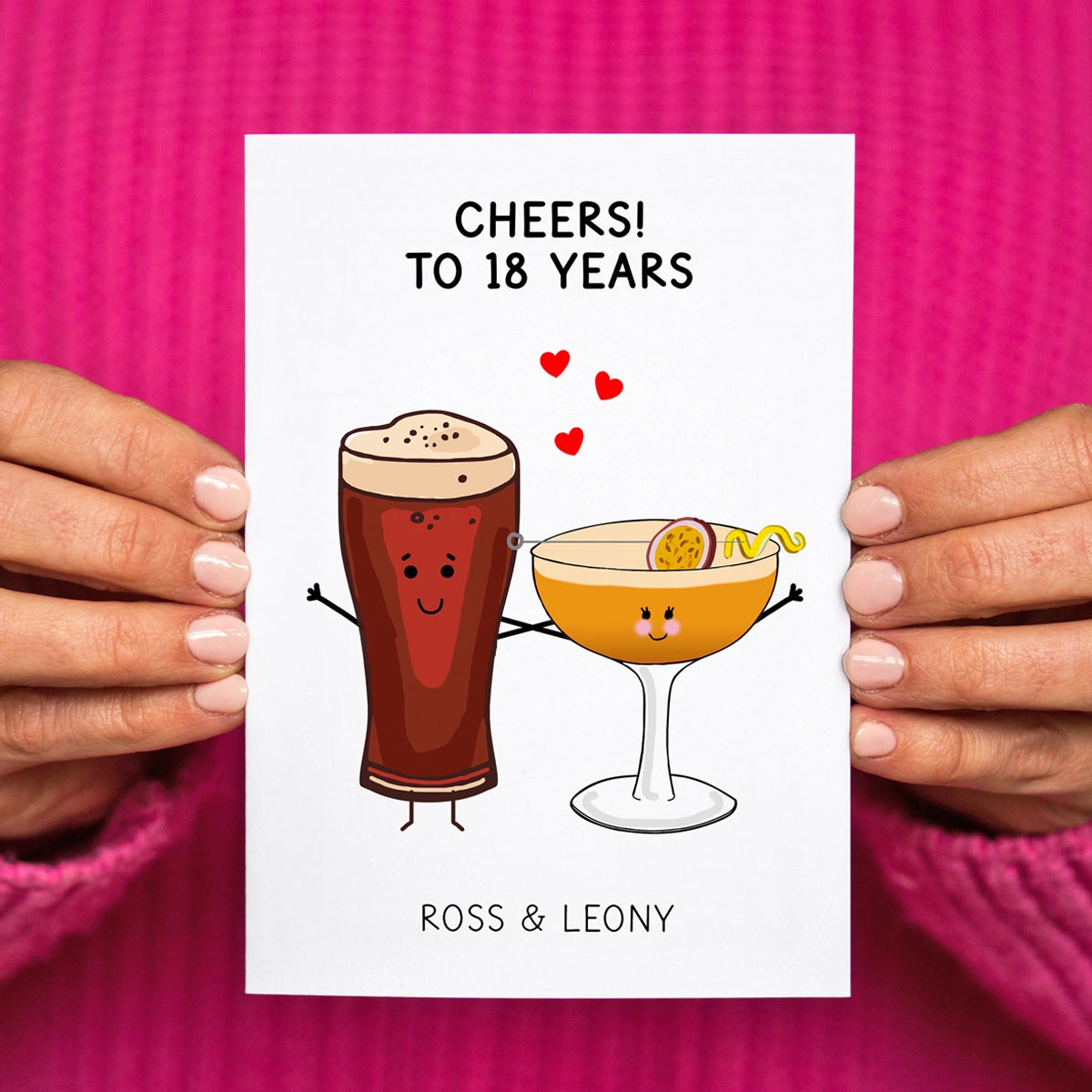 Greeting card with cartoon drinks and 'Cheers! To 18 Years' text held by hands against a pink background