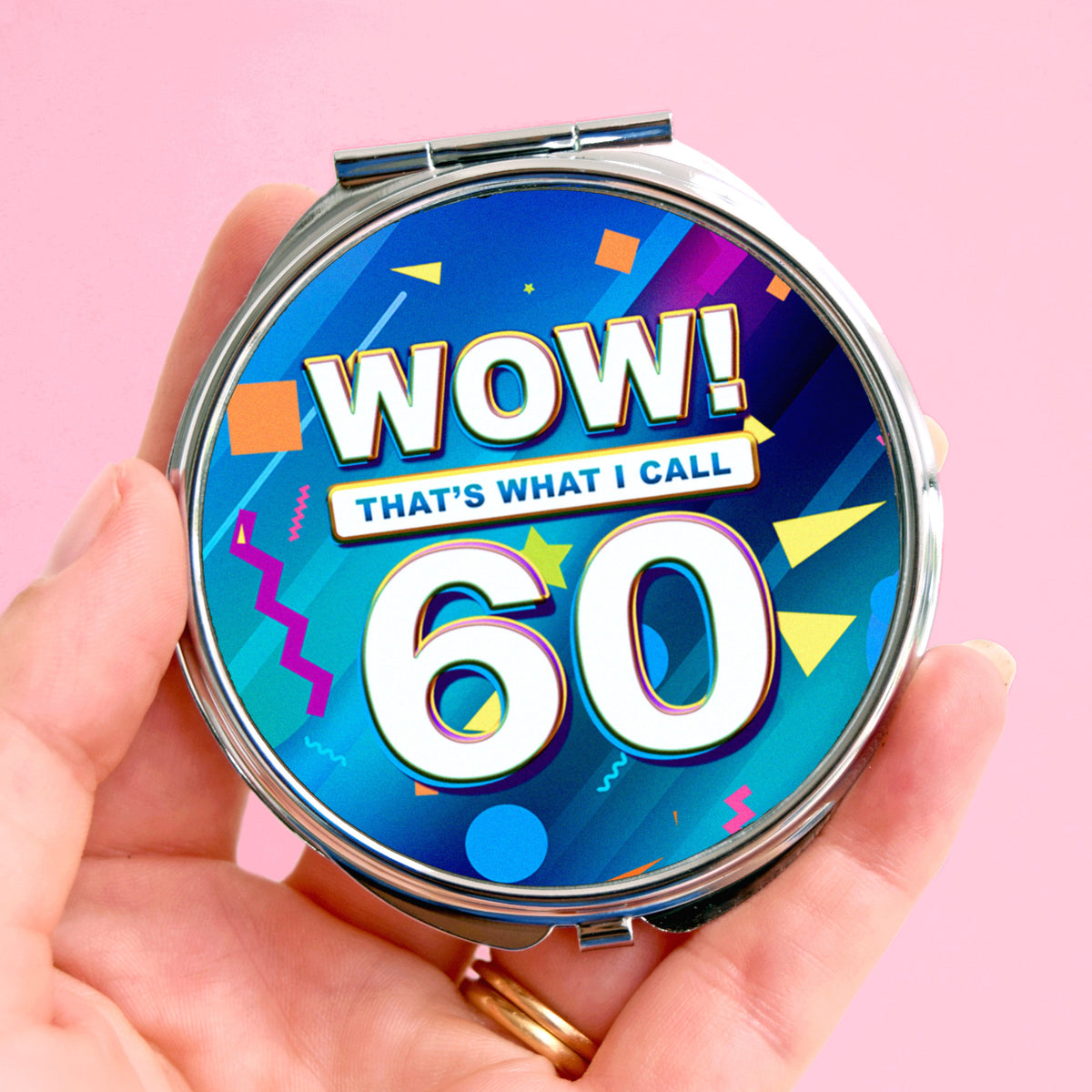 A hand holding a round compact mirror with a colorful retro 1960s-style graphic that reads 'WOW! THAT'S WHAT I CALL 60' on a solid pink background.