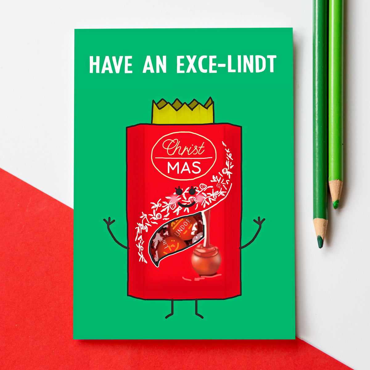 A green greeting card with a red illustrated Lindt chocolate box character and the text 'HAVE AN EXCE-LINDT Christmas' is shown on a white background with red corner shapes and two green pencils.