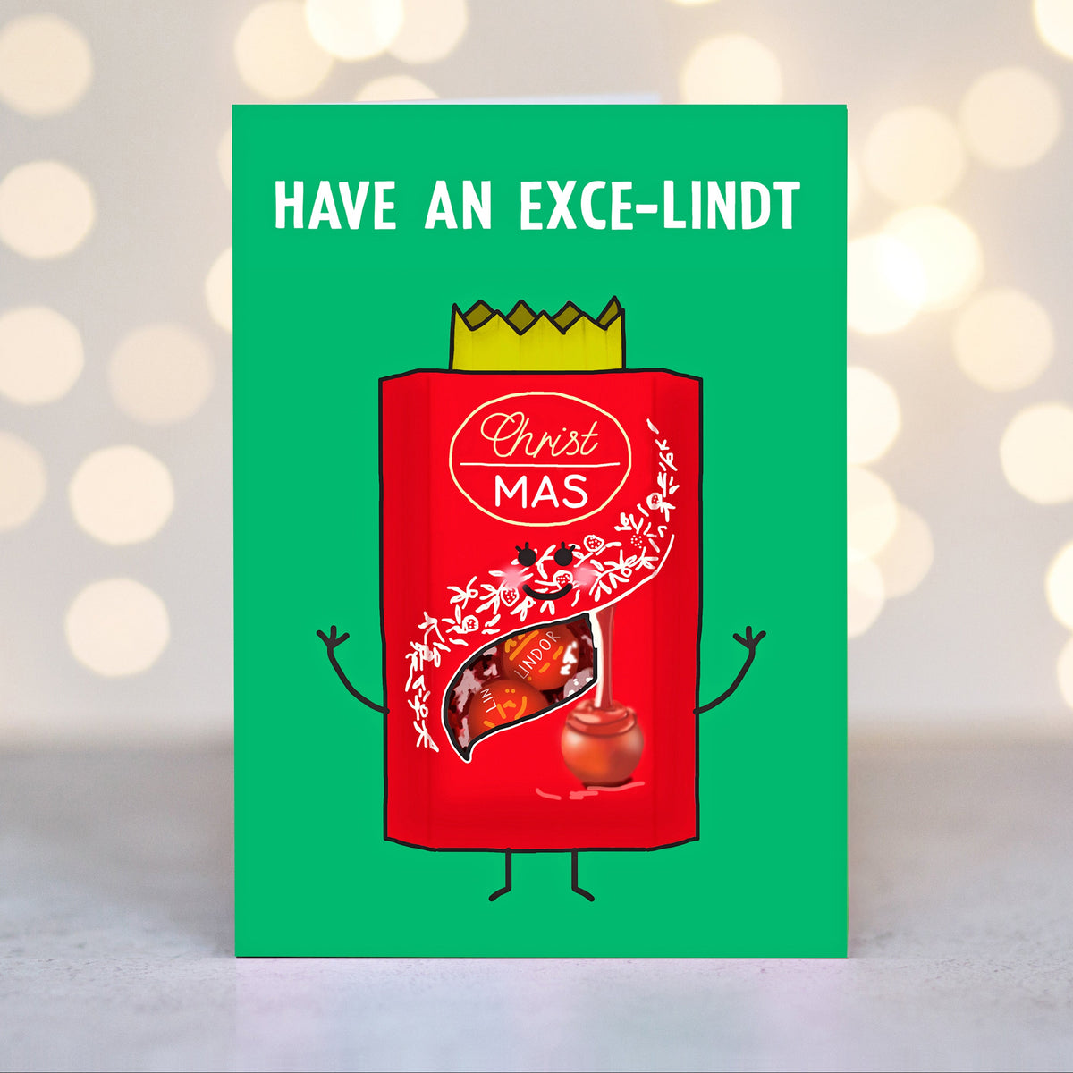A green greeting card featuring an illustration of a red box of Lindt chocolates with the text 'HAVE AN EXCE-LINDT CHIRSTMAS' at the top, photographed against a softly blurred warm bokeh background.
