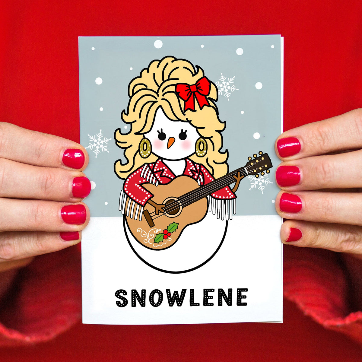 A person holds a Christmas card featuring a cartoon snowman dressed as Dolly Parton playing a guitar, with the word "SNOWLENE" printed below. The background is a bright red fabric, and only the person's hands and torso are visible.