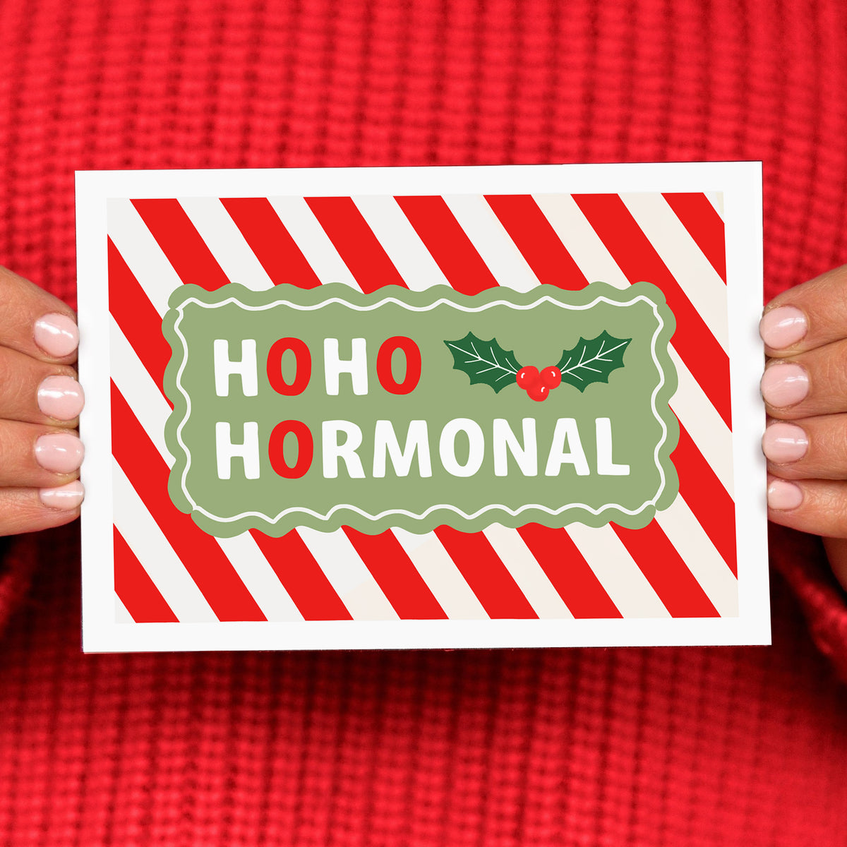 Card with 'HOHO HORMONAL' text and holly design held by hands against a red background