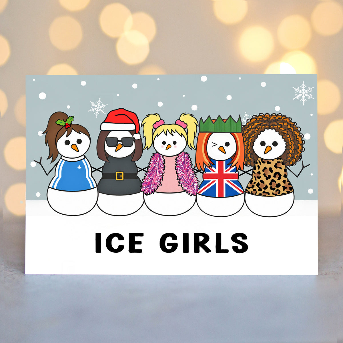 A Christmas card featuring a cartoon illustration of five snowmen dressed  in iconic Spice Girls outfits, with the text 'ICE GIRLS' printed below. The card is displayed against a blurred warm bokeh background.