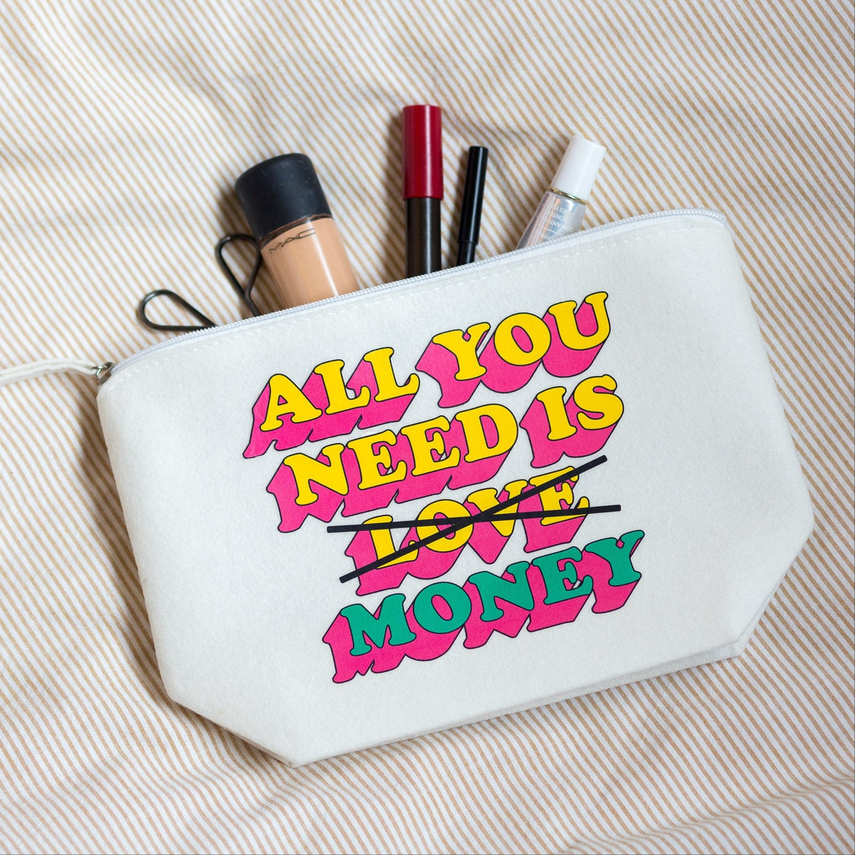 A white canvas cosmetic pouch with a colorful graphic reading 'ALL YOU NEED IS LOVE MONEY' in pink, yellow, and green, with 'LOVE' crossed out in black. Makeup items are tucked into the top zipper, and the pouch is laid on a beige striped fabric background.