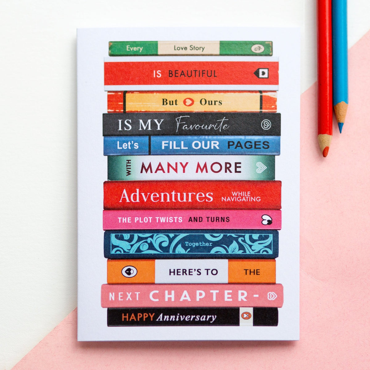 Card with book stack design and anniversary message on a pink surface with a pencil.