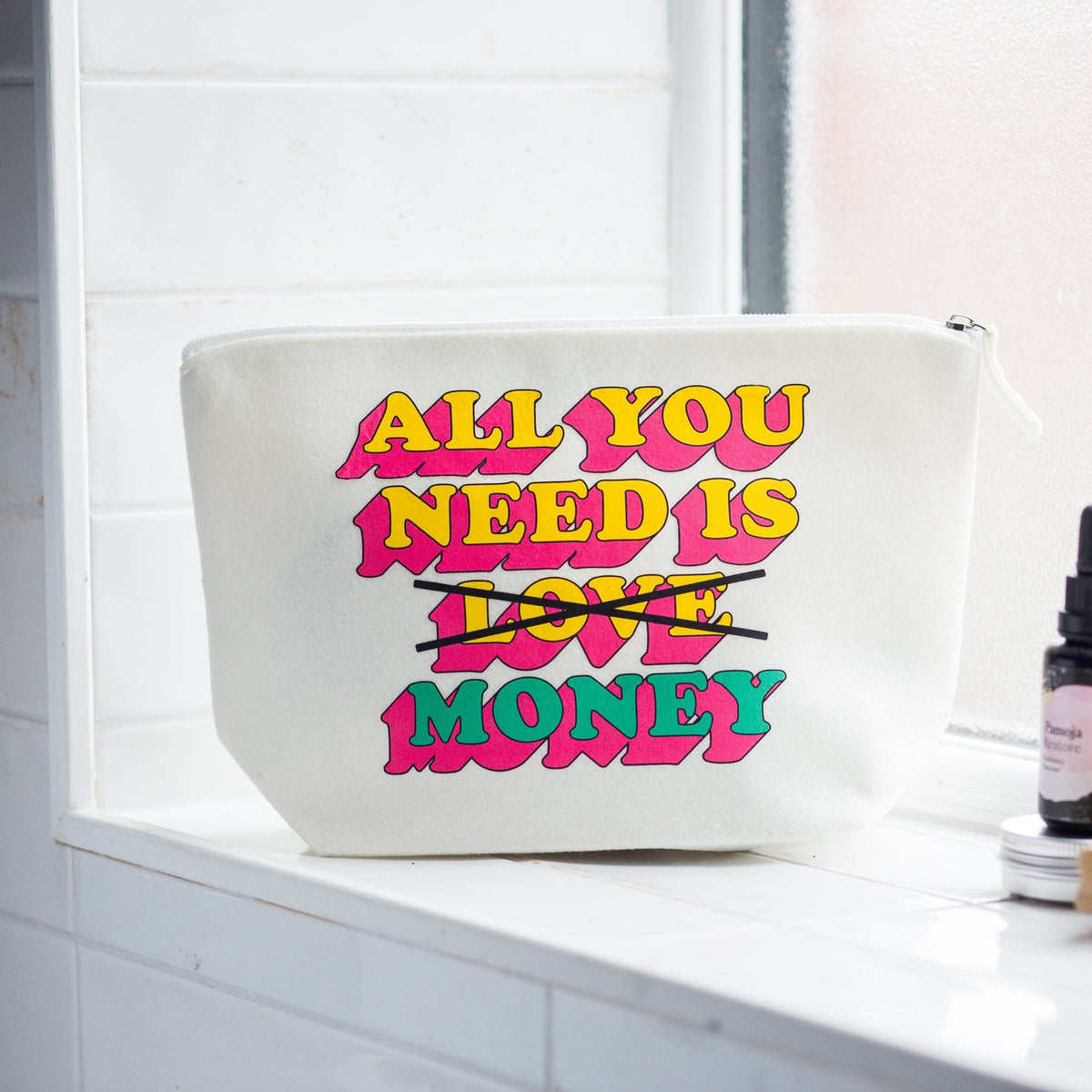 A white canvas cosmetic pouch with colorful retro text reading 'ALL YOU NEED IS LOVE MONEY' crossed out in black, sitting on a white tiled windowsill next to a small bottle.
