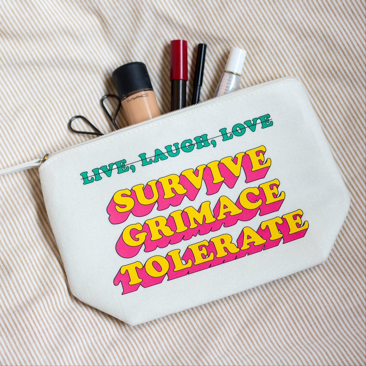 A white cosmetic pouch with colorful retro text reading 'LIVE, LAUGH, LOVE SURVIVE GRIMACE TOLERATE' lies on a beige striped fabric background, with makeup items peeking out from the top.