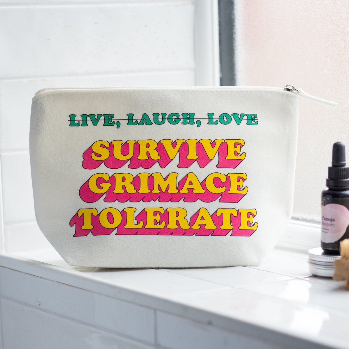 A white canvas cosmetic pouch with colorful retro text reading 'LIVE, LAUGH, LOVE SURVIVE GRIMACE TOLERATE' sits on a white tiled windowsill next to a small dropper bottle.