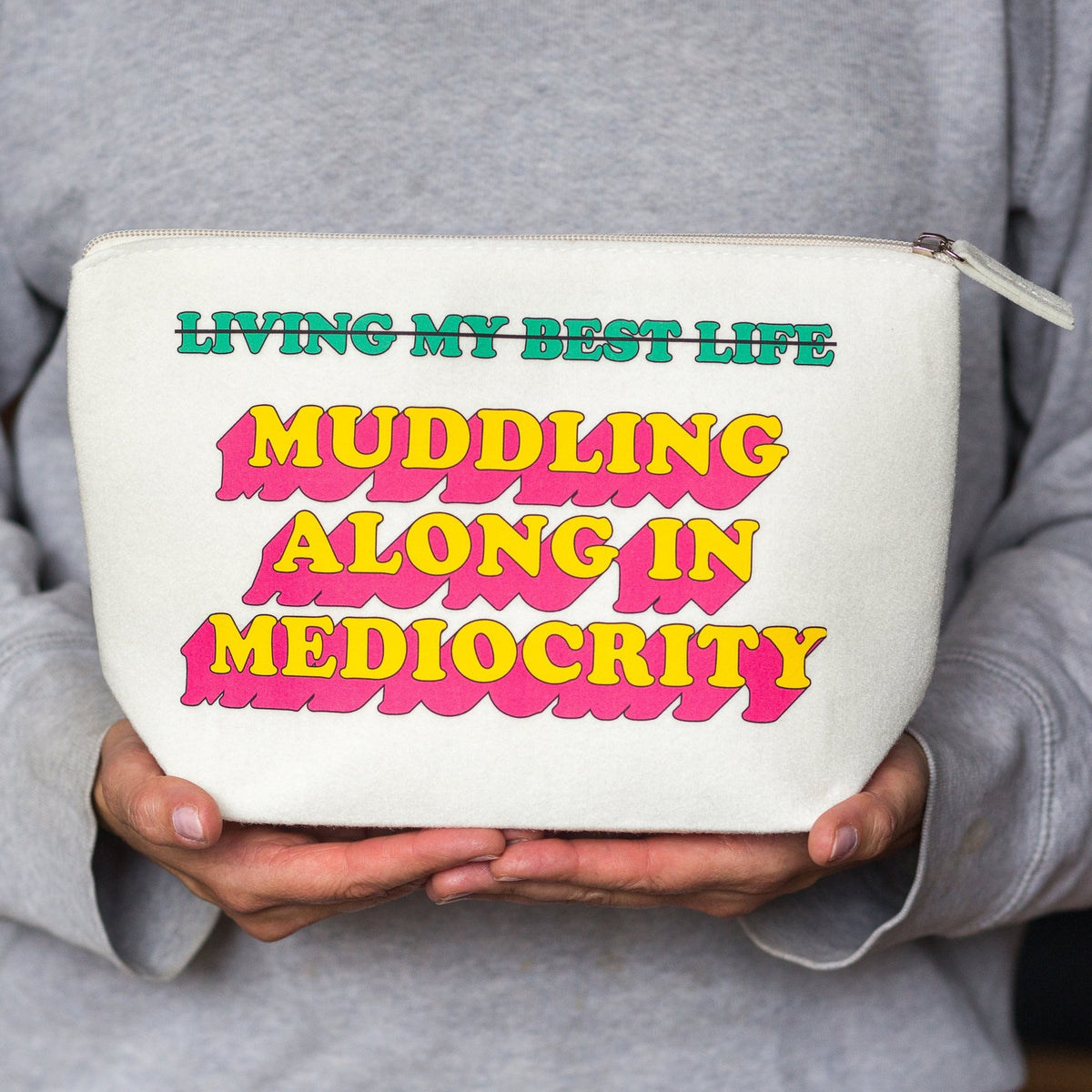A person in a gray sweatshirt holds a white zippered pouch with bold retro-style text reading 'LIVING MY BEST LIFE MUDDLING ALONG IN MEDIOCRITY' in green, pink, and yellow.