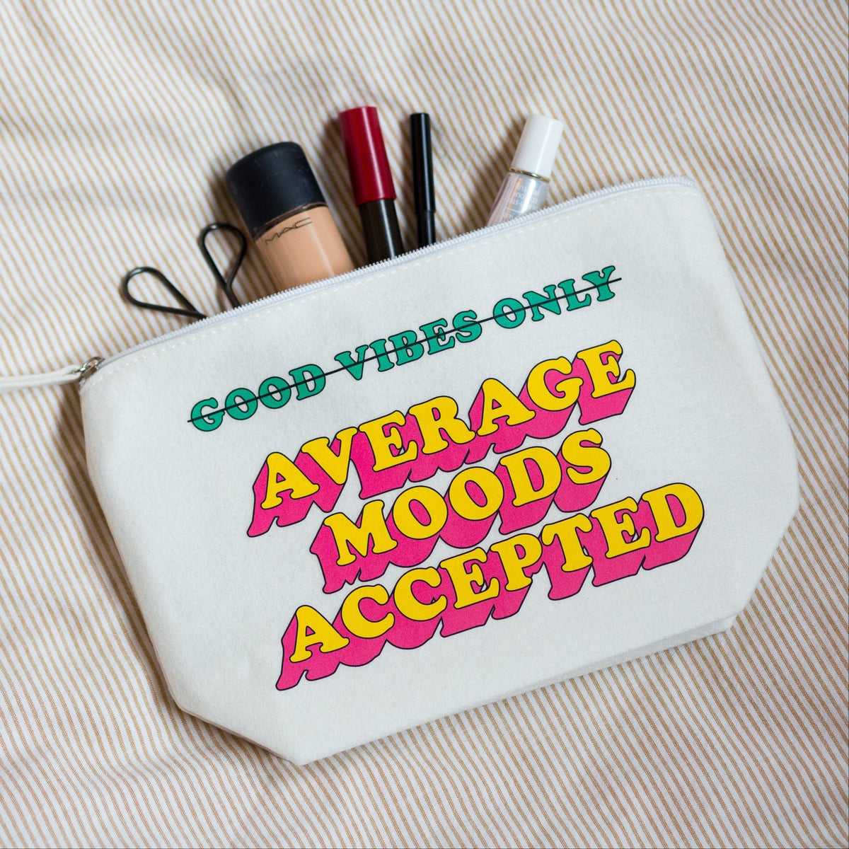 A cream-colored canvas cosmetic pouch with colorful retro text reading 'GOOD VIBES ONLY AVERAGE MOODS ACCEPTED', shown on a beige striped fabric background with makeup items peeking out from the top.