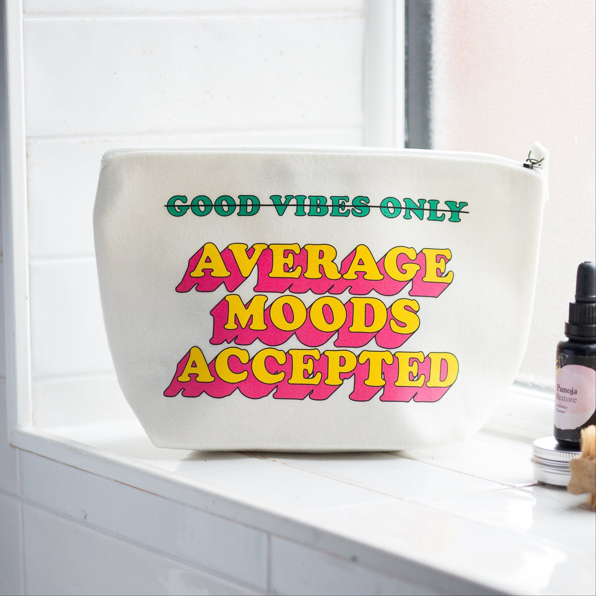 A white cosmetic pouch with colorful retro text reading 'GOOD VIBES ONLY AVERAGE MOODS ACCEPTED' sits on a white tiled windowsill next to a small bottle.