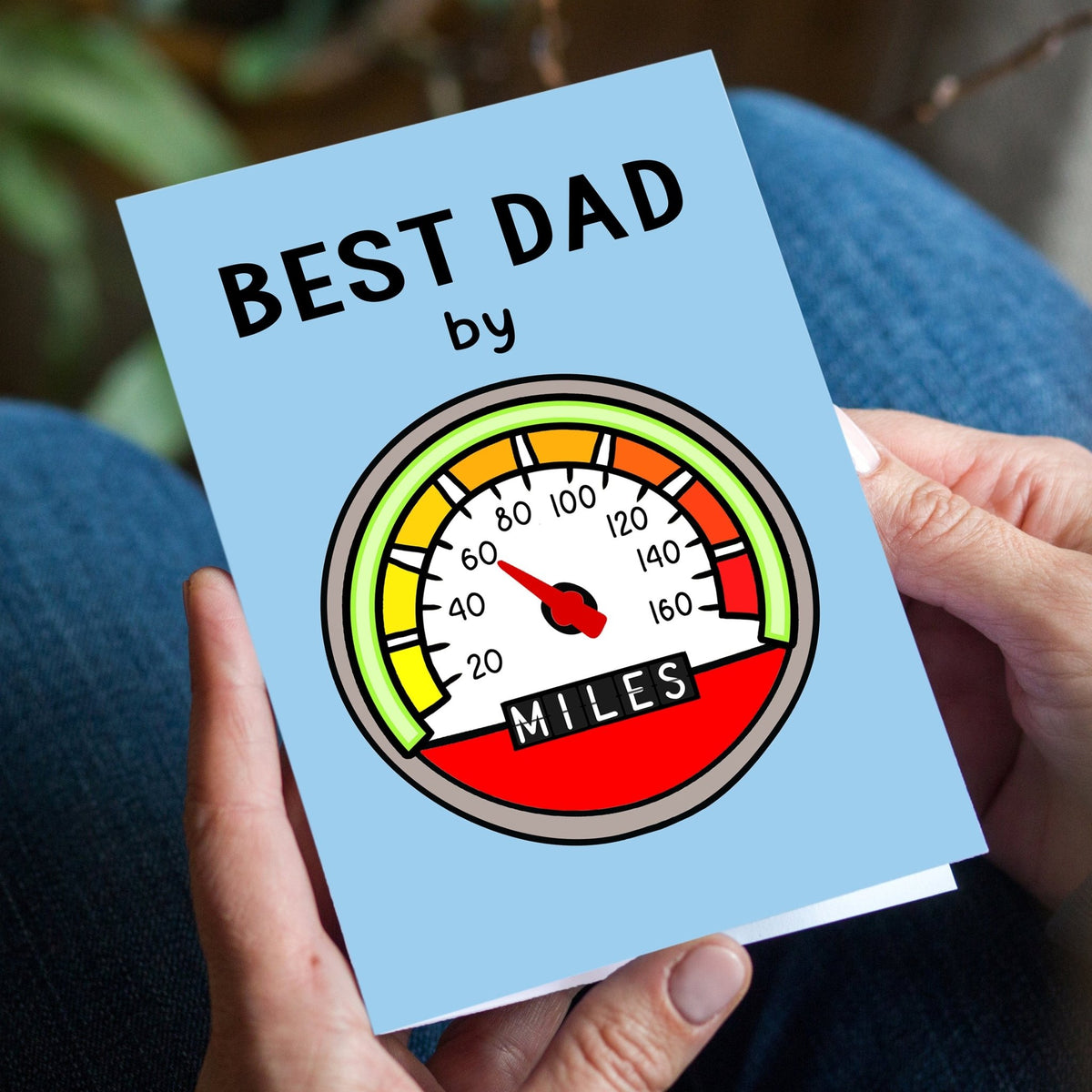 Card with 'Best Dad by Miles' text and speedometer graphic held by a person.