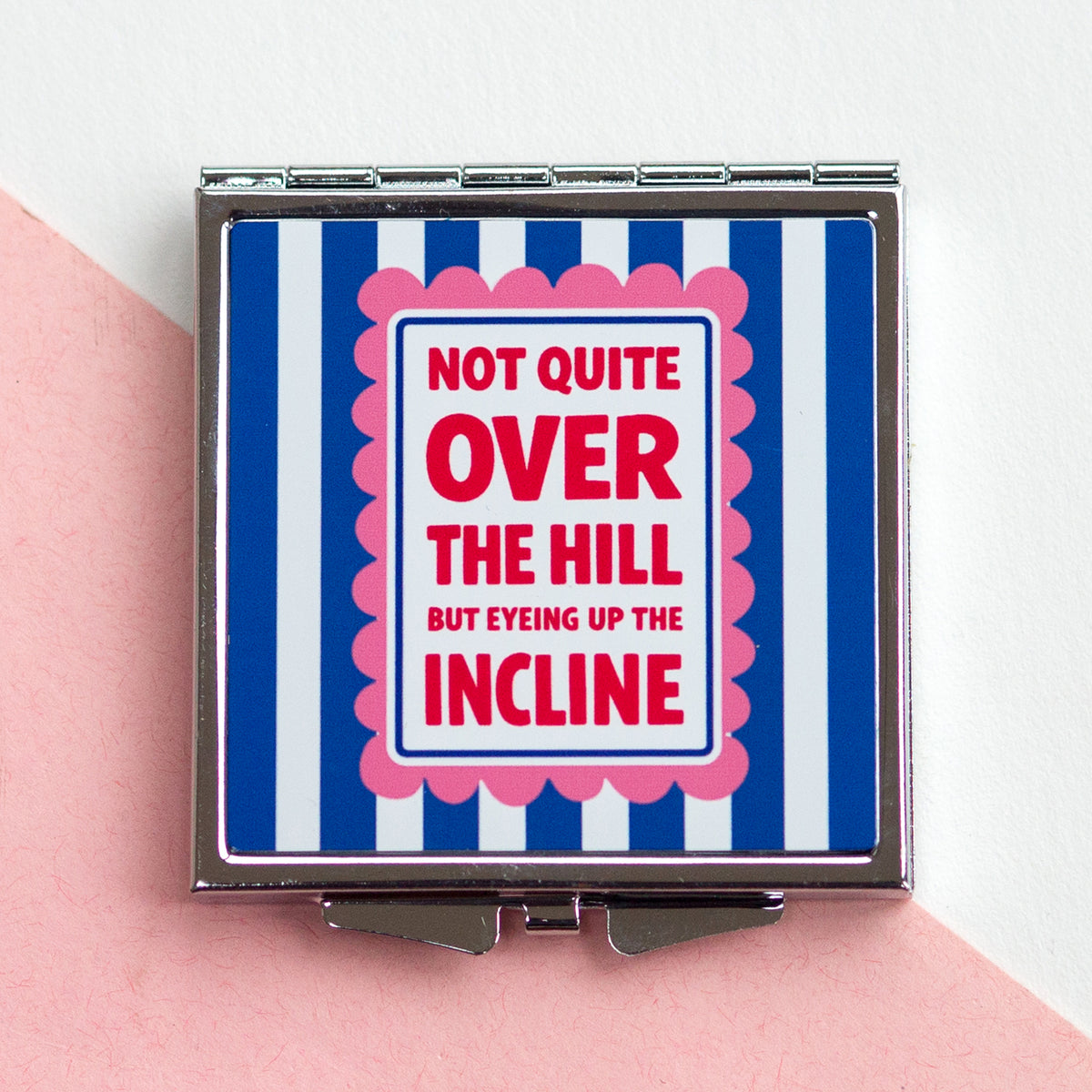 Compact mirror with blue and white striped design and humorous text on a pink surface.
