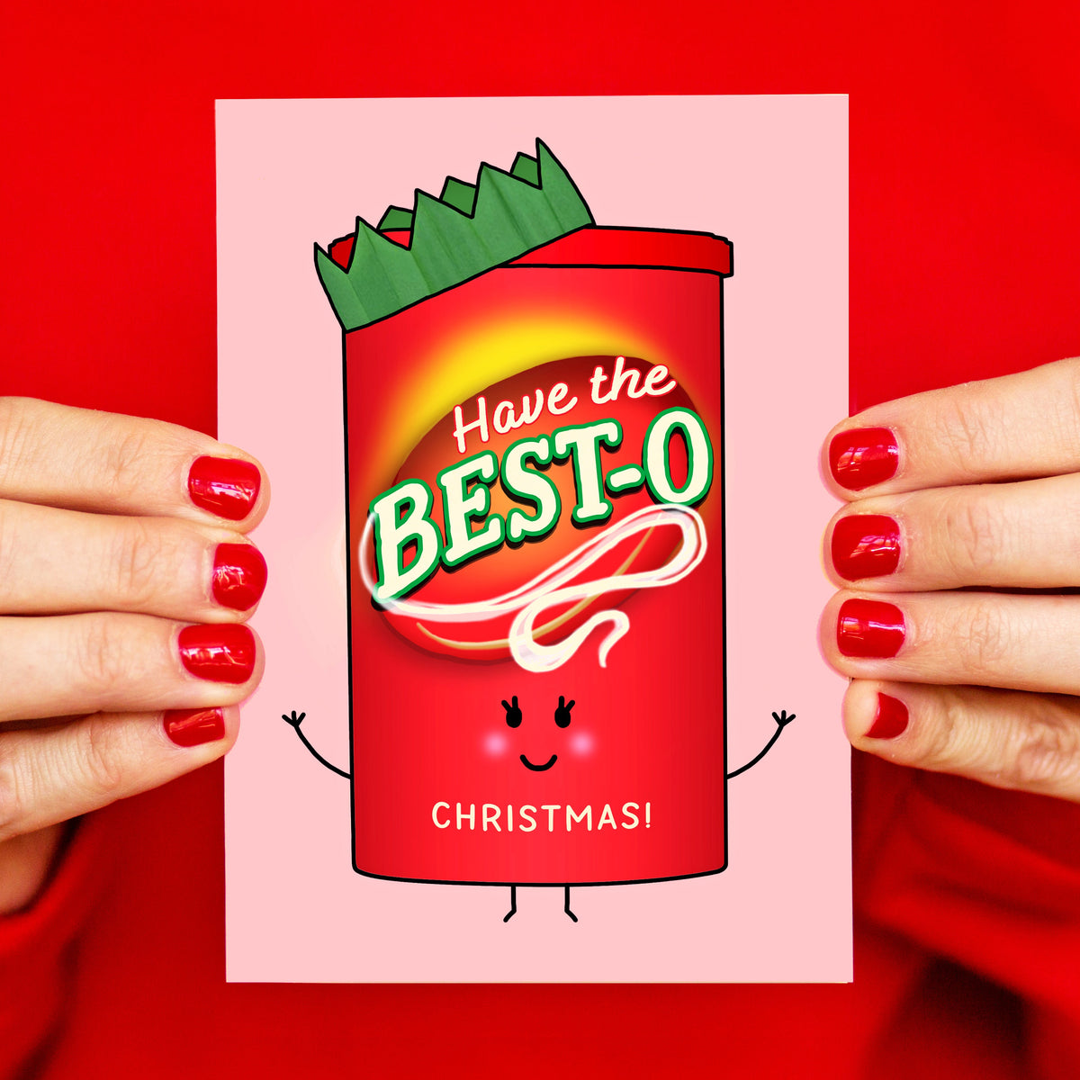 A person with red-painted nails holds a pink greeting card featuring a cartoon red candy canister with the text 'Have the BEST-O CHRISTMAS!'. The background is a solid bright red.