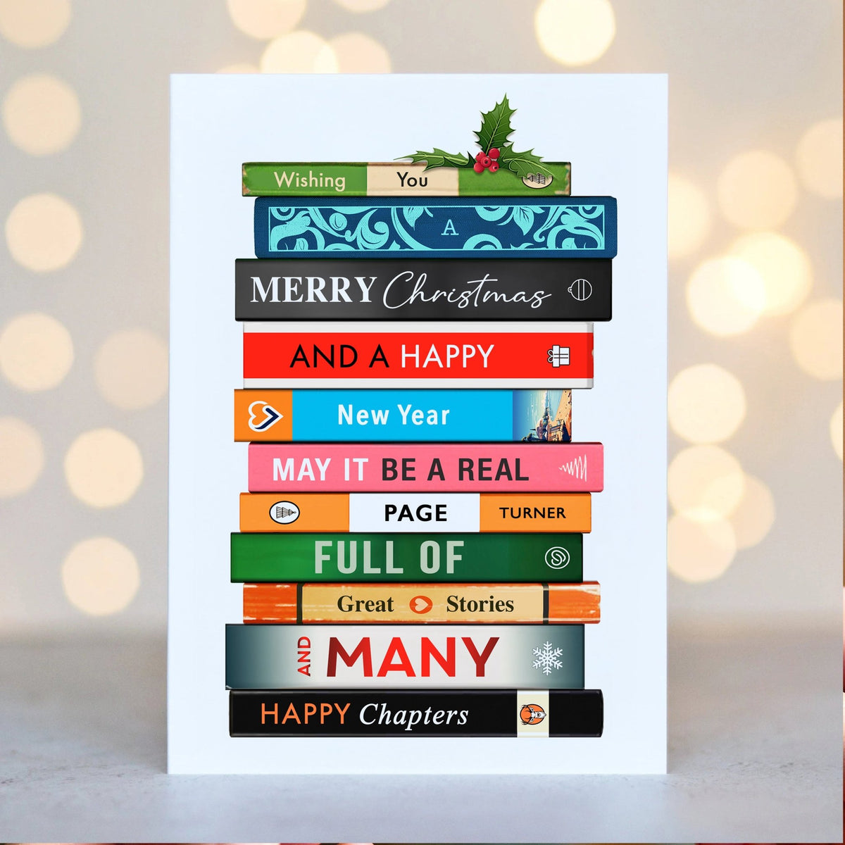 A white rectangular greeting card standing upright on a light surface, printed with a stack of colorful books and holiday text including 'Wishing You', 'MERRY Christmas', 'AND A HAPPY New Year', 'MAY IT BE A REAL Happy Chapter', and 'HAPPY Chapters'. The background is softly blurred with warm bokeh lights.