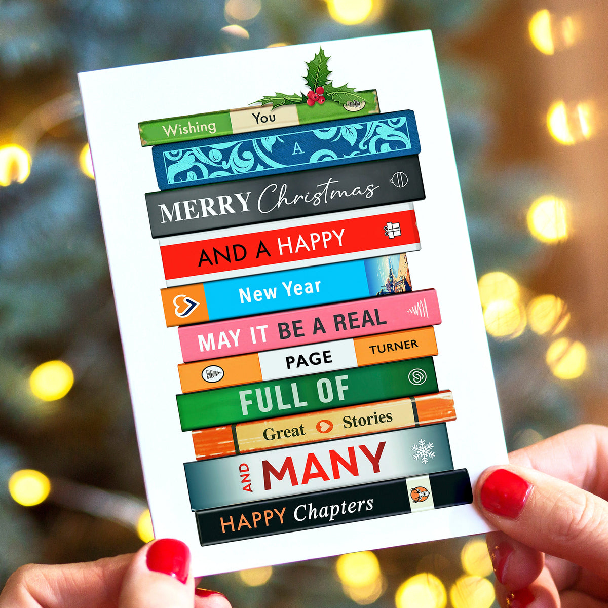 A person holds a white Christmas greeting card featuring colorful book spines stacked to spell out holiday greetings. The background shows a blurred Christmas tree and warm bokeh lights, making this a Lifestyle shot.