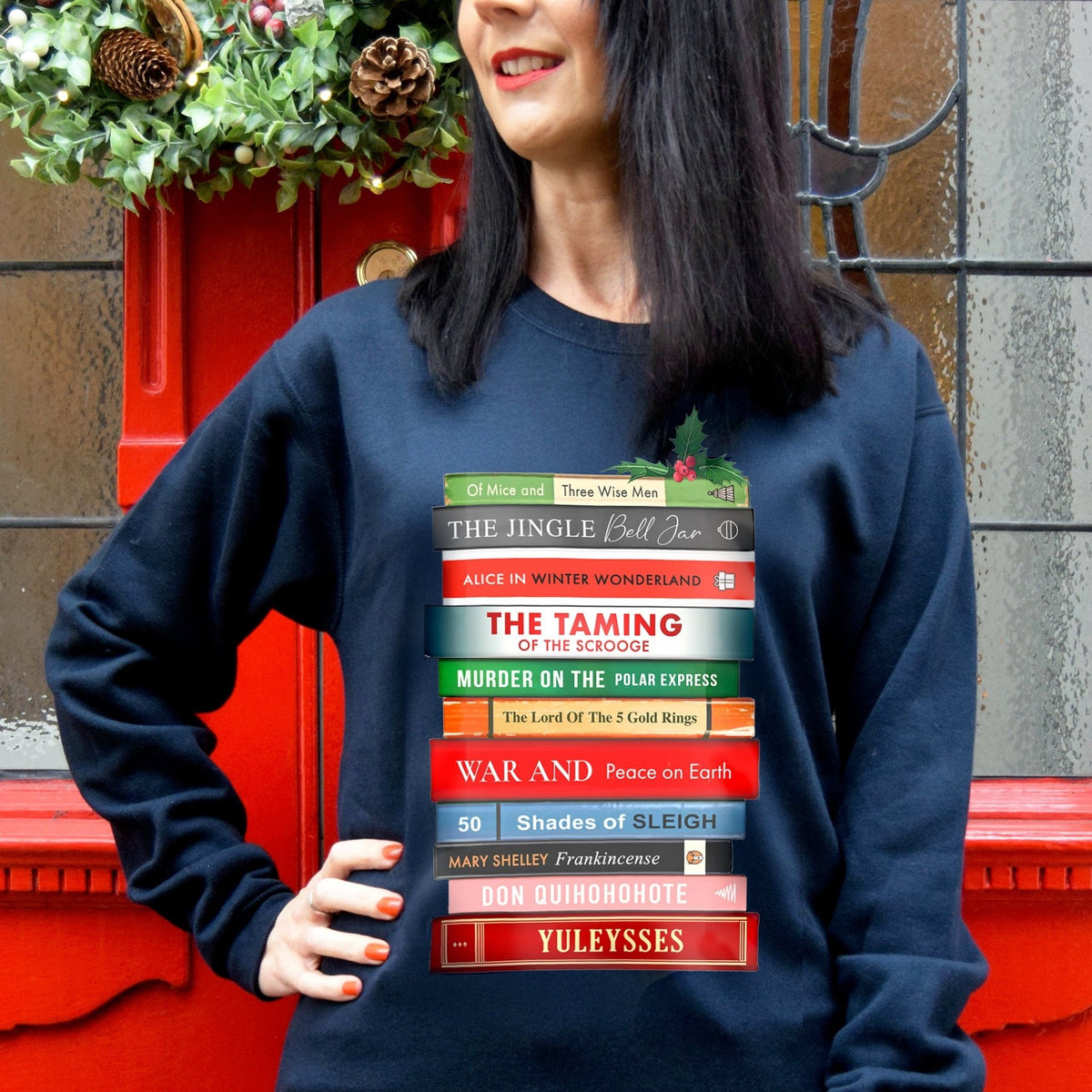 Person wearing a navy Christmas sweatshirt with a stack of books featuring festive puns on classic book book titles in front of a red door.