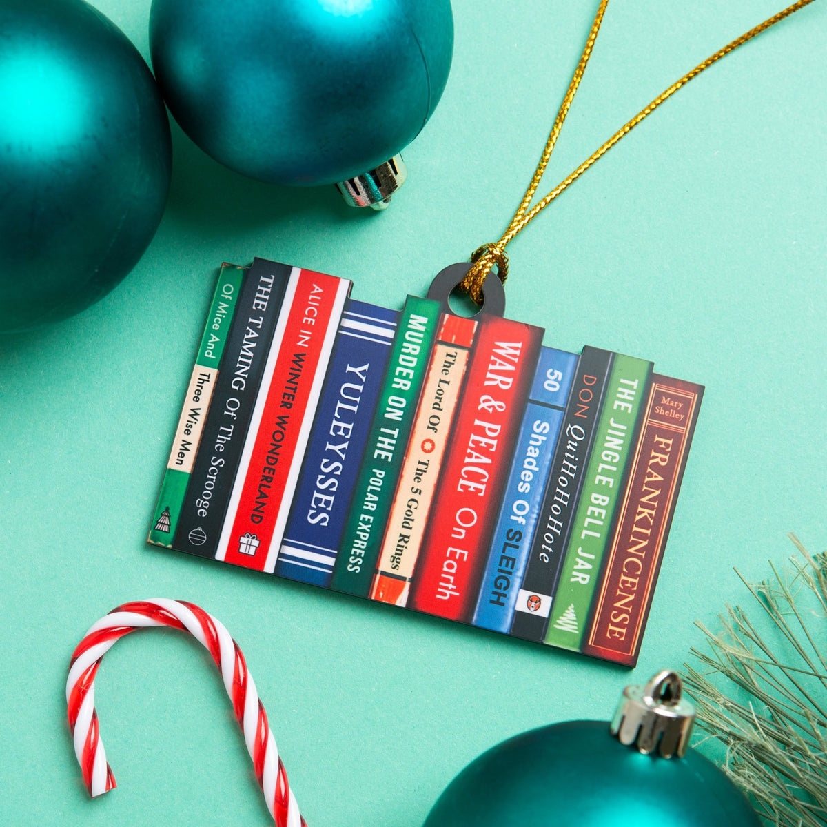 A Christmas ornament shaped like a row of colorful book spines hangs from a gold string on a mint green background, with teal ornaments, a candy cane, and pine branches around it.