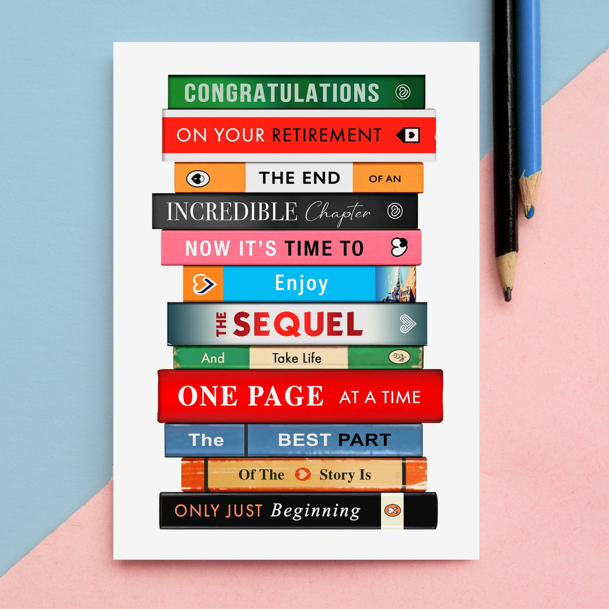 A white retirement card featuring a stacked row of colourful book spines with a book themed retirement message, photographed on a pastel blue and pink background with two pencils at the top right.