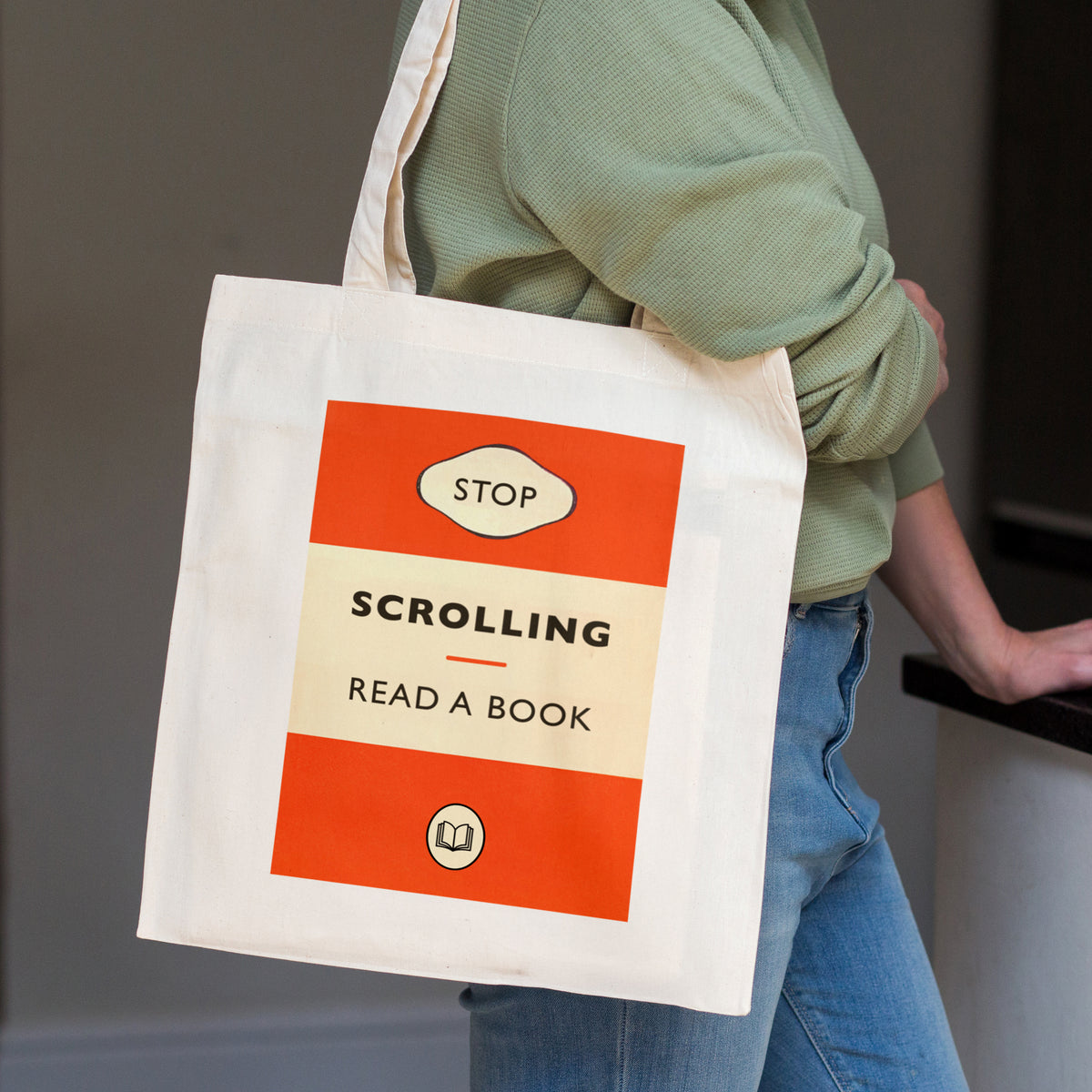 A person wearing a sage green long-sleeve shirt and blue jeans carries a cream canvas tote bag with a retro book cover design that reads 'STOP SCROLLING READ A BOOK'.