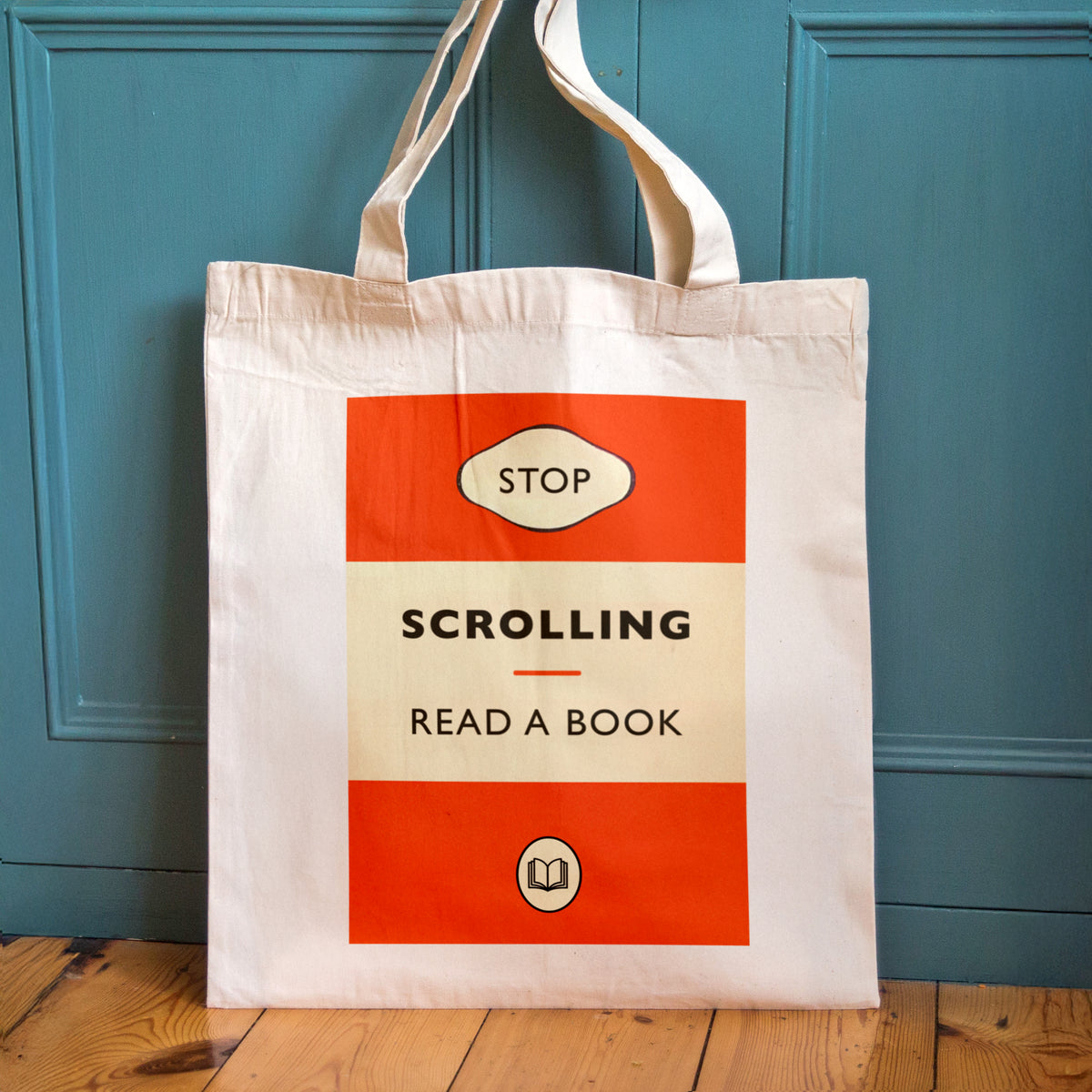 A cream canvas tote bag with a retro-style book cover graphic that reads 'STOP SCROLLING READ A BOOK' in orange and beige, standing against a teal paneled door on a wooden floor.