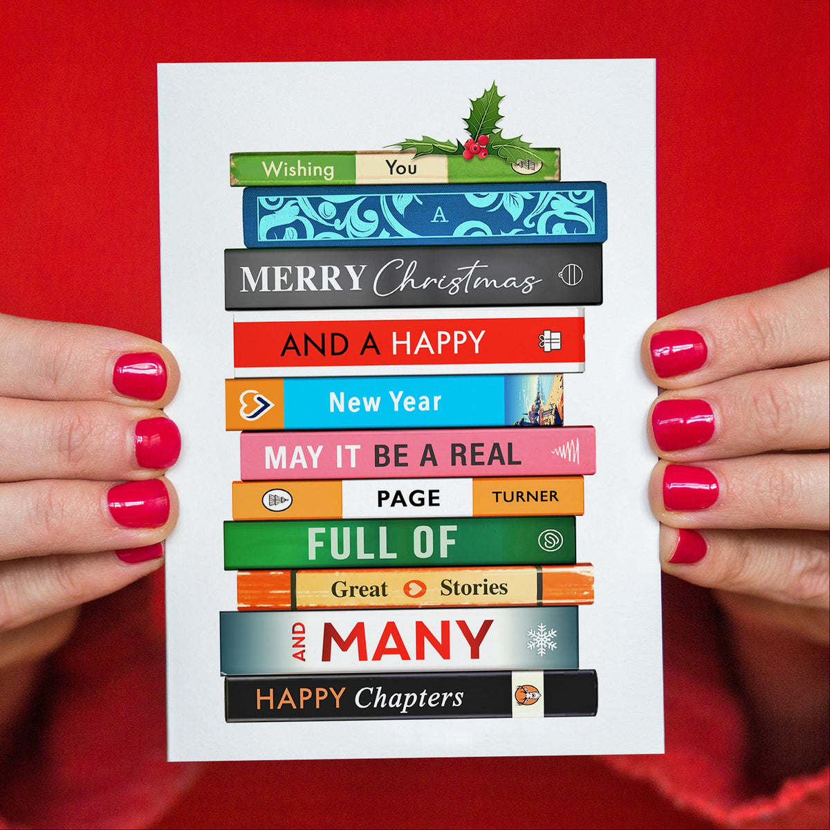 A white Christmas greeting card held by two hands with bright pink fingernails against a solid red background. The card features an illustration of stacked book spines in various colors with festive holiday and New Year themed text like 'Wishing You', 'Merry Christmas', 'New Year', and 'Happy Chapters'.