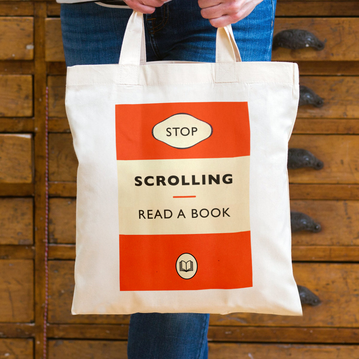 A person holding a cream canvas tote bag with a retro book cover design that reads 'STOP SCROLLING READ A BOOK' in orange and black, set against a background of wooden bookshelves.
