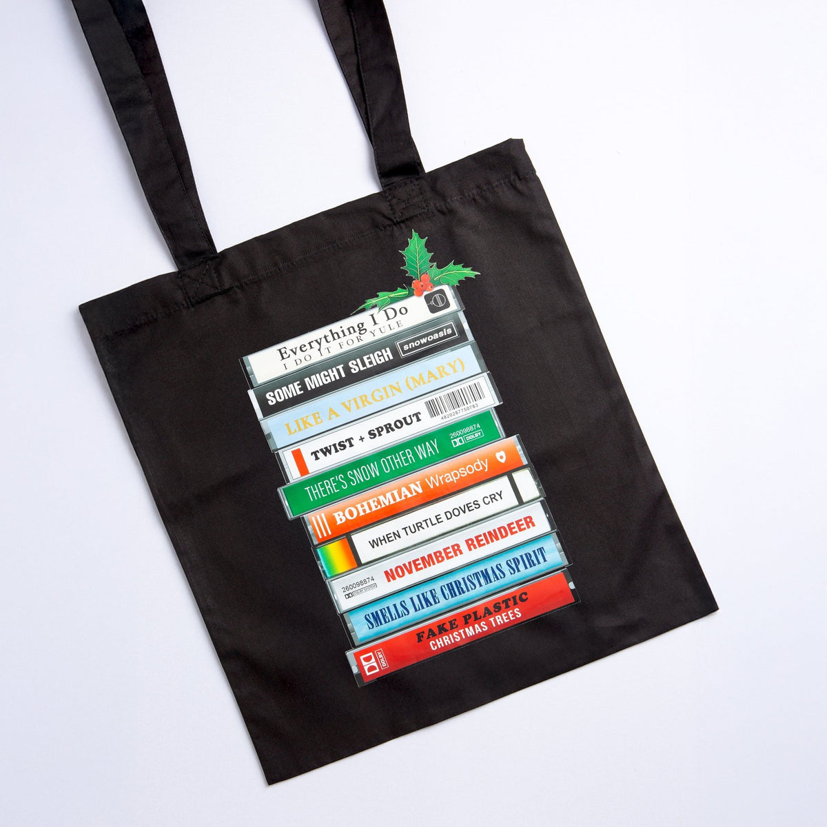 A black canvas tote bag with two shoulder straps, featuring a printed graphic of stacked cassette tapes with festive puns on classic song titles, photographed on a clean white background.