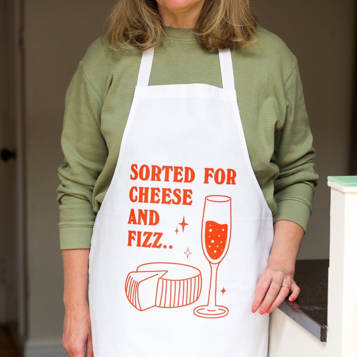 A woman wearing a white apron with orange text that reads 'SORTED FOR CHEESE AND FIZZ.