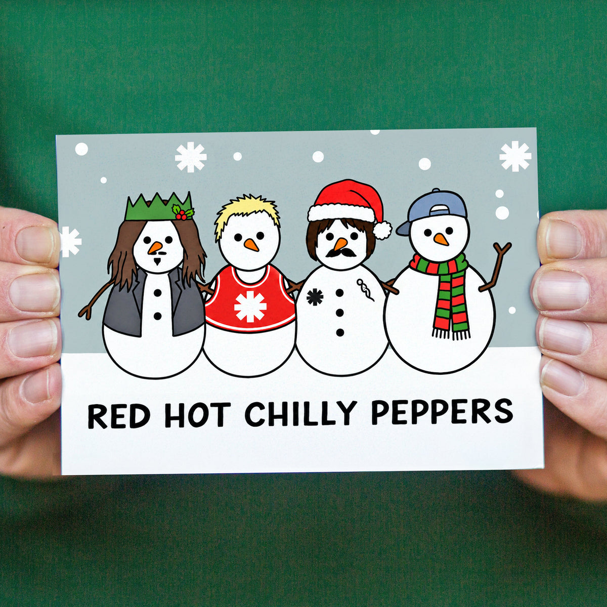 A hand holds a rectangular novelty card featuring four cartoon snowmen with winter clothing and the text 'RED HOT CHILLY PEPPERS' on a green background.
