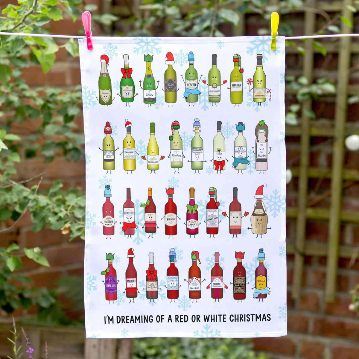 A white tea towel with a colorful illustration of Christmas wine bottles and the text 'I'M DREAMING OF A RED OR WHITE CHRISTMAS' hanging from a clothesline outdoors with pink and yellow clips, against a background of green foliage and a brick wall.