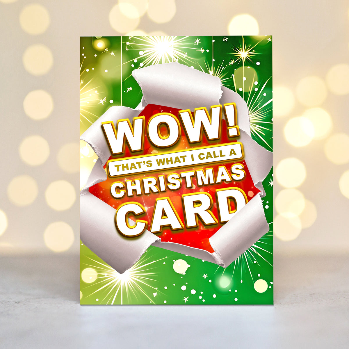 A Christmas greeting card with a green and red festive design, featuring torn white paper edges and the text 'WOW! THAT'S WHAT I CALL A CHRISTMAS CARD' in bold white letters with gold outlines, set against a blurred warm bokeh background.