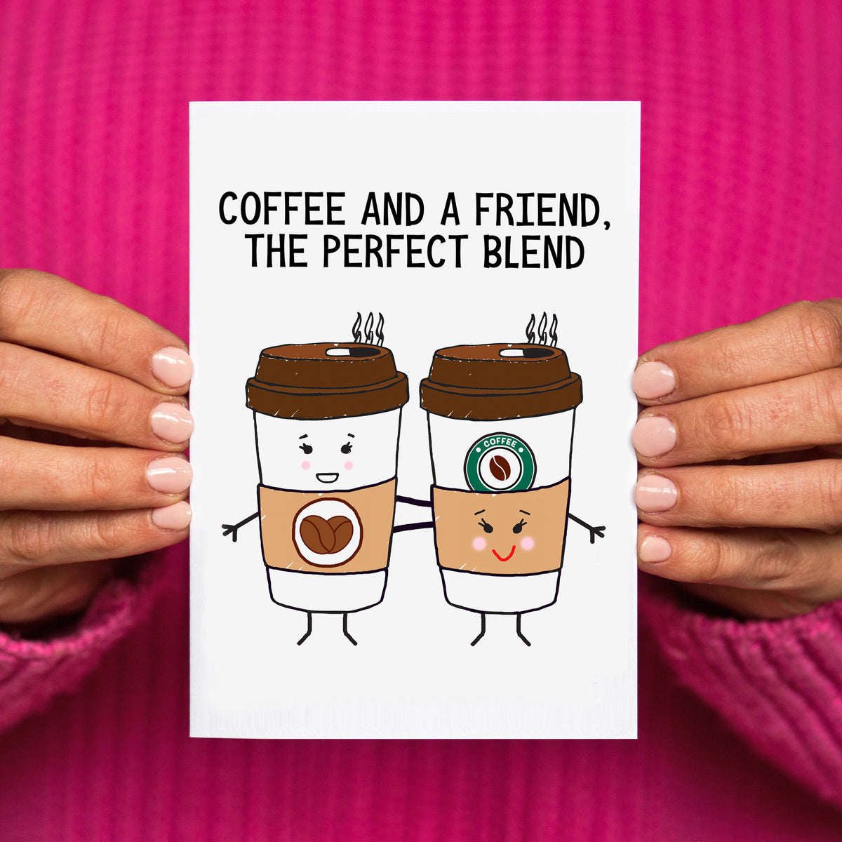 A white greeting card held by two hands against a bright pink knit sweater. The card features the text 'COFFEE AND A FRIEND, THE PERFECT BLEND' above two cartoon takeaway coffee cups with faces and arms.