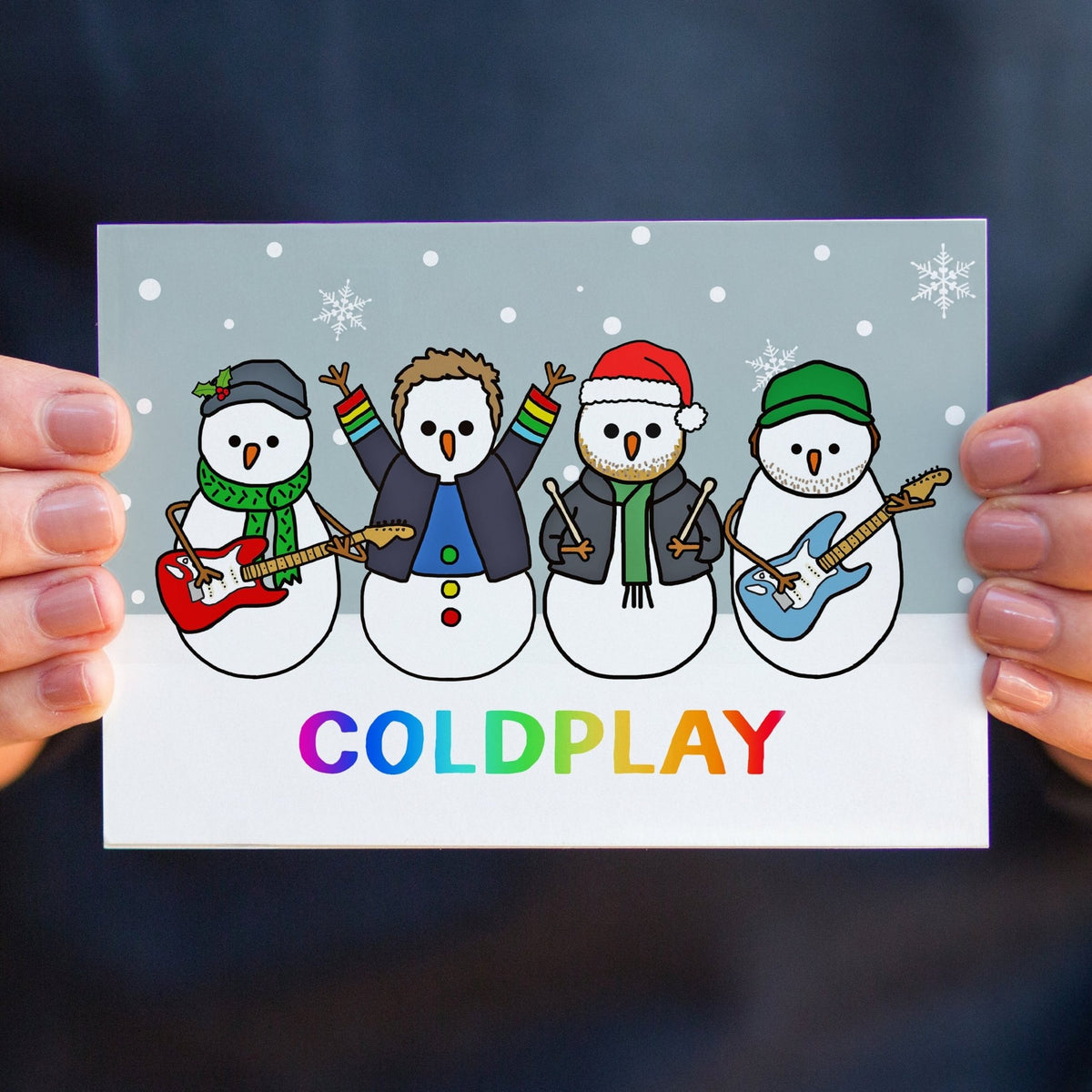 A person holds a Christmas card featuring four cartoon snowmen playing guitars with the word COLDPLAY in rainbow letters. The card is shown against a dark navy background, with the person's hands visible on both sides.