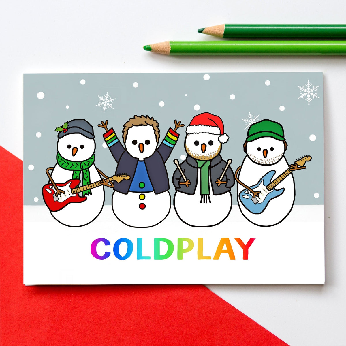 A Christmas card featuring four cartoon snowmen playing guitars with the word 'COLDPLAY' in rainbow letters at the bottom. The card is photographed on a white surface with red paper in the corner and two green pencils at the top.