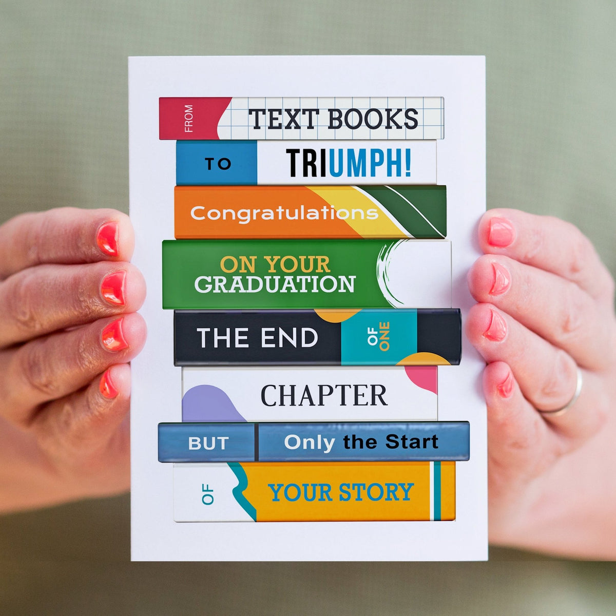Graduation card with colorful text books design held by hands against a green background