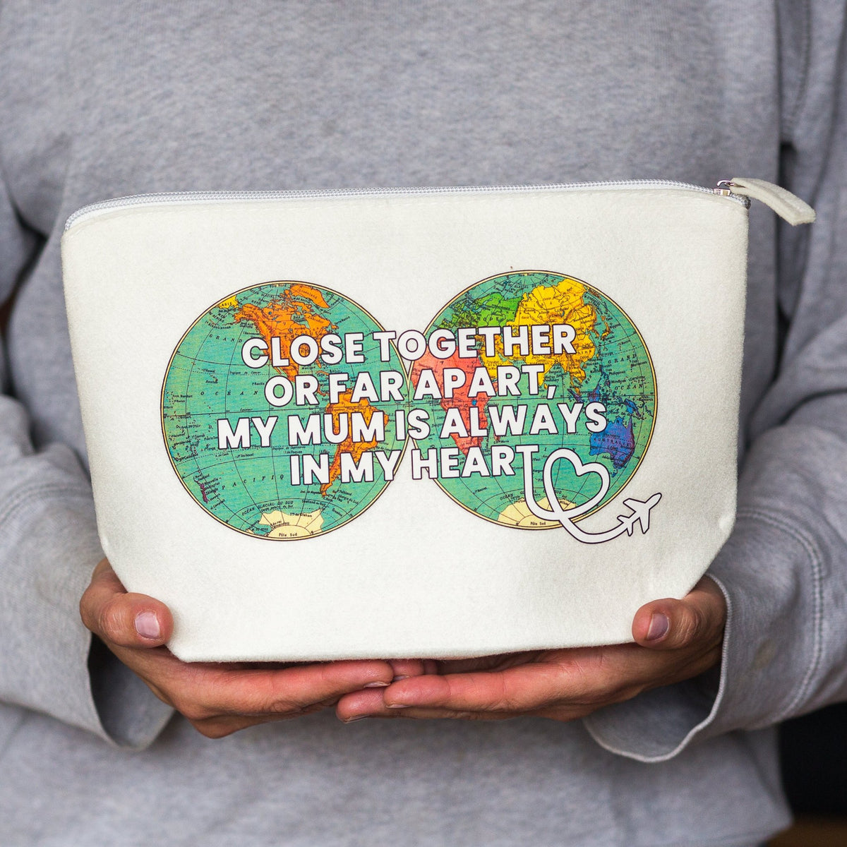 A person in a gray hoodie holds a cream-colored zippered pouch. The pouch features a graphic of two matching globe designs in green, orange, and blue, joined by a heart shape, with the text 'CLOSE TOGETHER OR FAR APART, MY MUM IS ALWAYS IN MY HEART' in white.