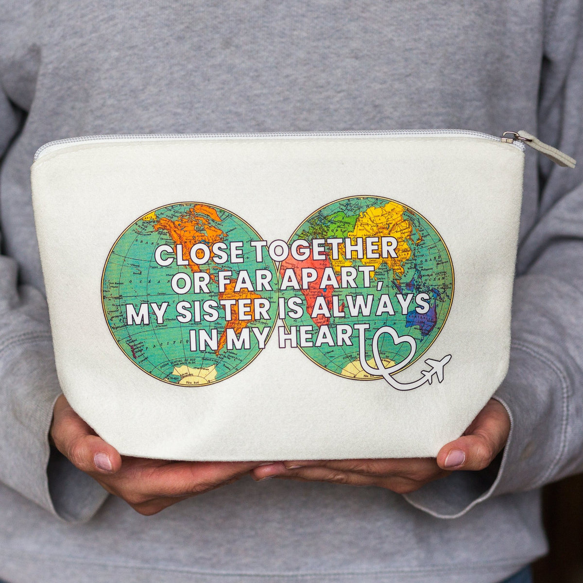 A person in a gray sweatshirt holds a cream-colored canvas zippered pouch. The pouch features a graphic of two green map circles side-by-side with the text 'CLOSE TOGETHER OR FAR APART, MY SISTER IS ALWAYS IN MY HEART' and a small white heart with an airplane.