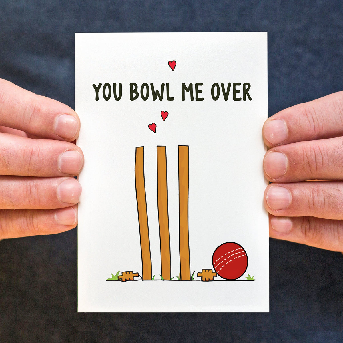 Card with cricket-themed design and text 'You Bowl Me Over' held by two hands against a dark background.