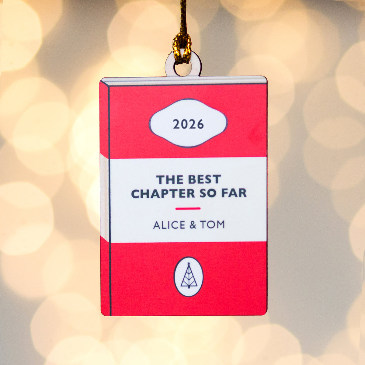 A red and white book-shaped ornament hanging from a gold string against a warm bokeh background. The ornament reads '2026', 'THE BEST CHAPTER SO FAR', and 'ALICE & TOM'.