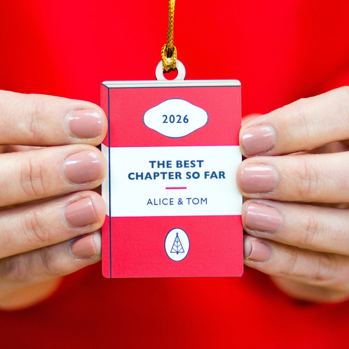 A person holds a red and white book-shaped ornament with a gold hanging string. The ornament reads '2026', 'THE BEST CHAPTER SO FAR', 'ALICE & TOM', and has a small tree logo near the bottom.