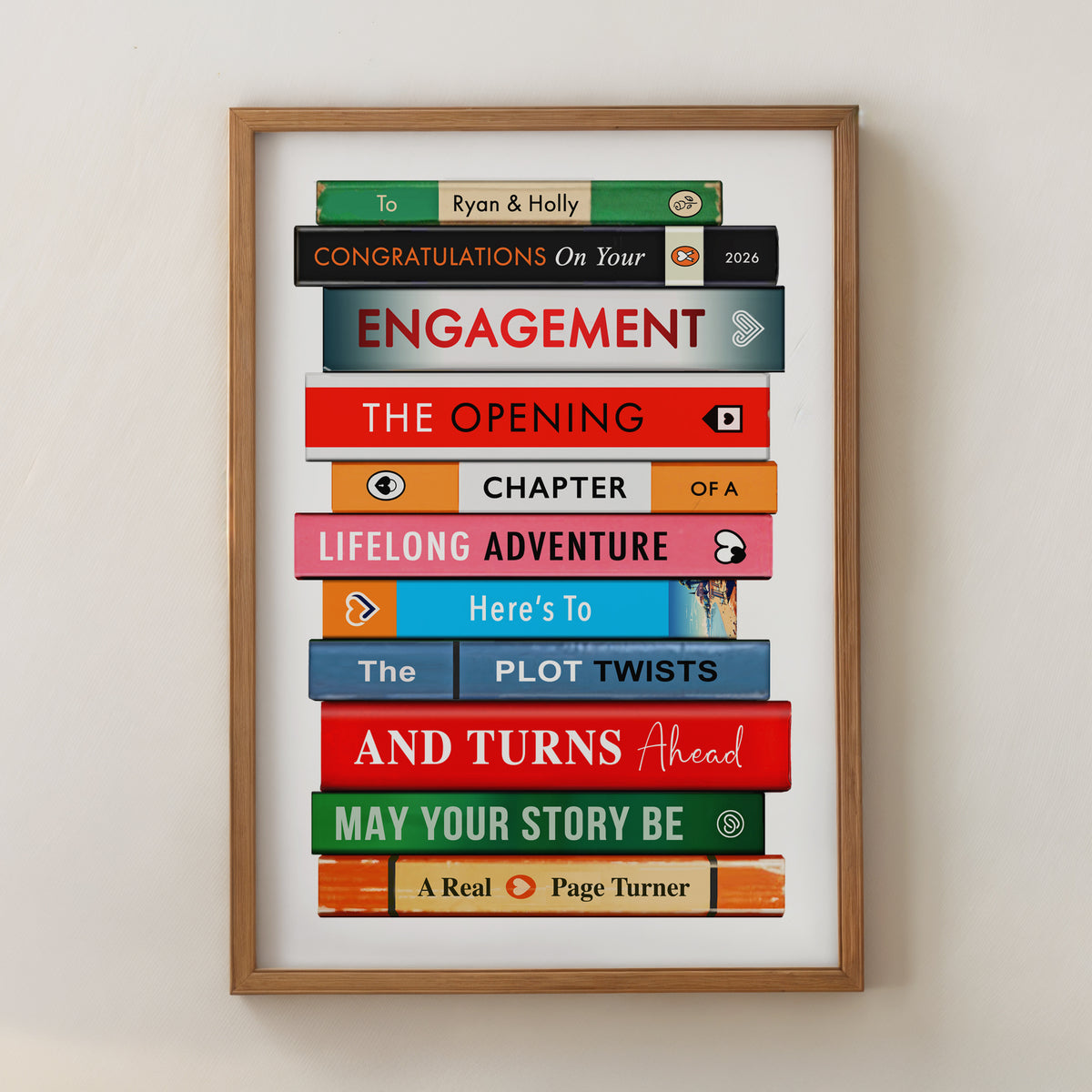 A framed personalised book stack wall art print with colorful book spines and text celebrating engagement, shown hanging on a light beige wall.