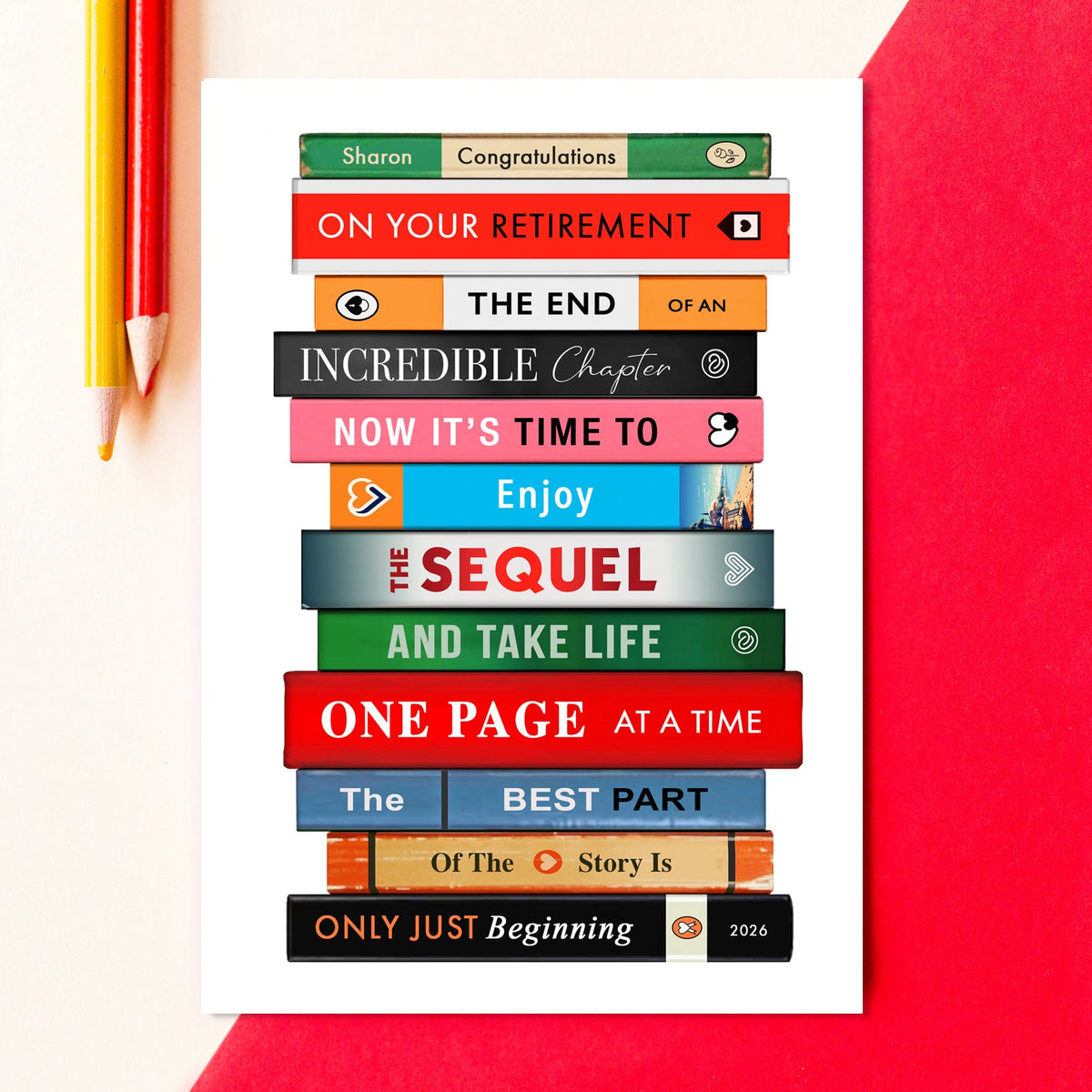 A square white art print featuring a stacked book design with various colorful book spines and text like 'Congratulations', 'On Your Retirement', and 'The Sequel and Take Life'. The print is styled on a white surface with a red paper corner and two colored pencils.