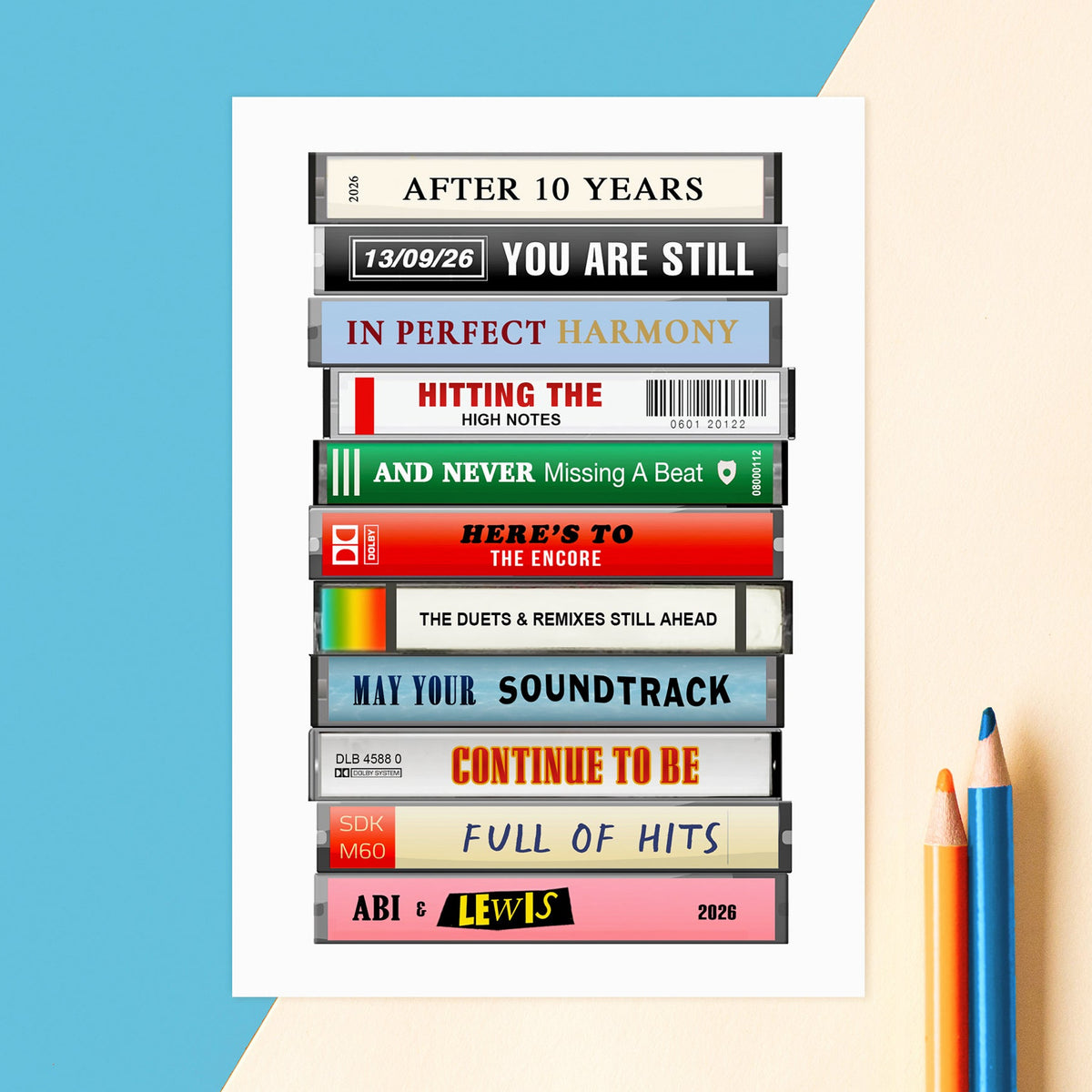 A vertical art print featuring a stacked arrangement of vintage cassette tapes with various song titles and text, presented on a white card against a diagonally split blue and beige background with colored pencils on the right.