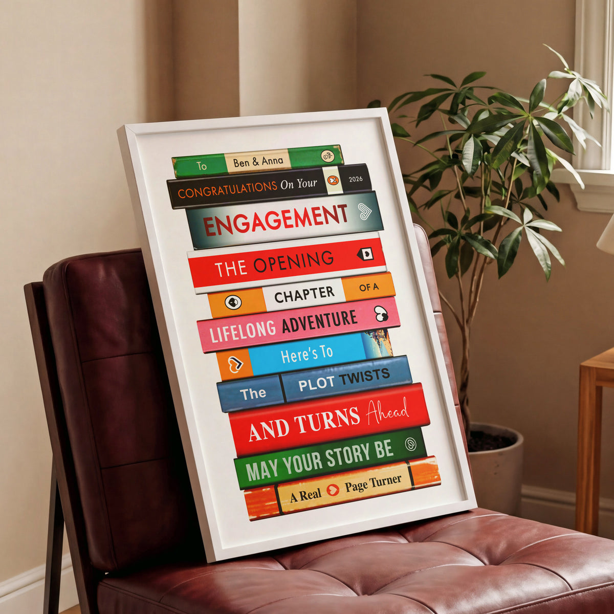 A framed personalised book-stack art print leaning against a brown leather chair in a living room setting next to a potted plant.