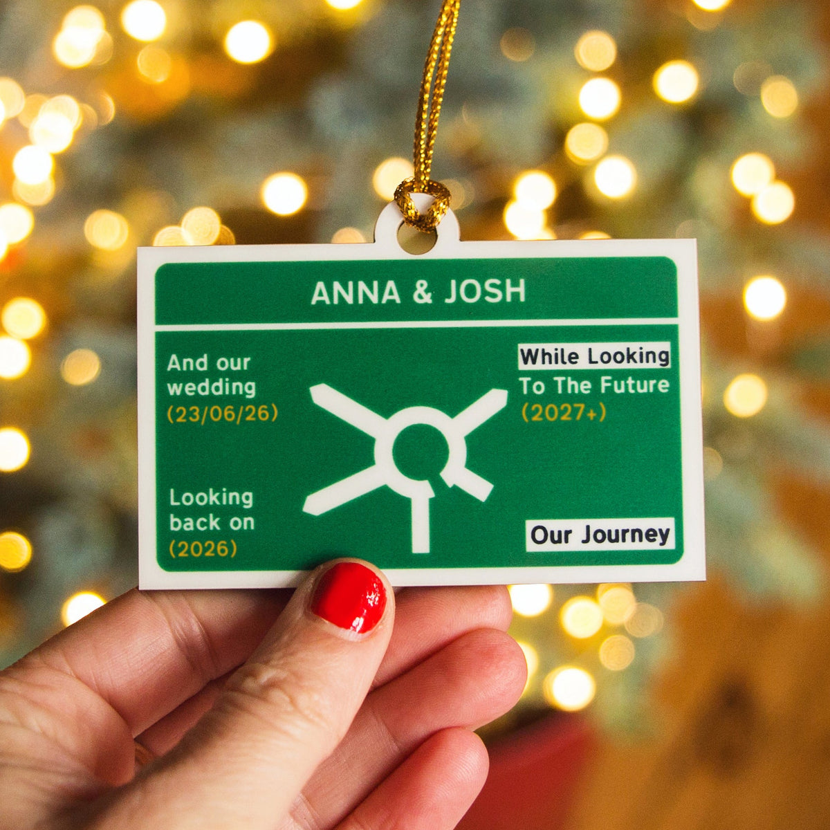 A personalised custom road sign Christmas ornament held by a hand with red nail polish, featuring custom wedding text and a white symbol, with a festive blurred background of warm bokeh lights.