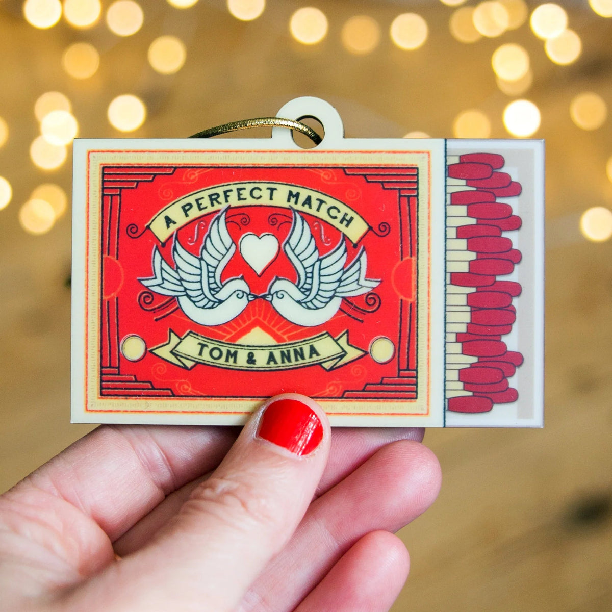 Matchbox labeled 'A Perfect Match Tom & Anna' held by a hand with red nail polish against a blurred light background.