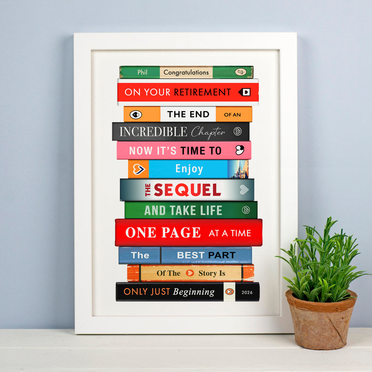 A framed personalized retirement poster styled on a white shelf next to a potted green plant. The poster features colorful book spines stacked vertically with humorous retirement-themed quotes like 'Congratulations', 'The End', and 'Now it's time to enjoy'.