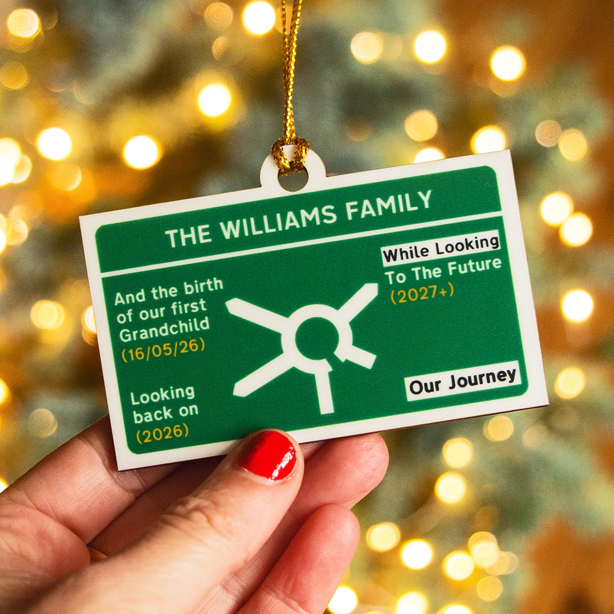 A hand with red nail polish holds a rectangular green and white personalised Christmas tree decoration designed to look like a motorway road sign with a gold hanging string. The ornament features the text 'THE WILLIAMS FAMILY' and custom milestone notes, with a blurred Christmas tree and warm bokeh lights in the background.
